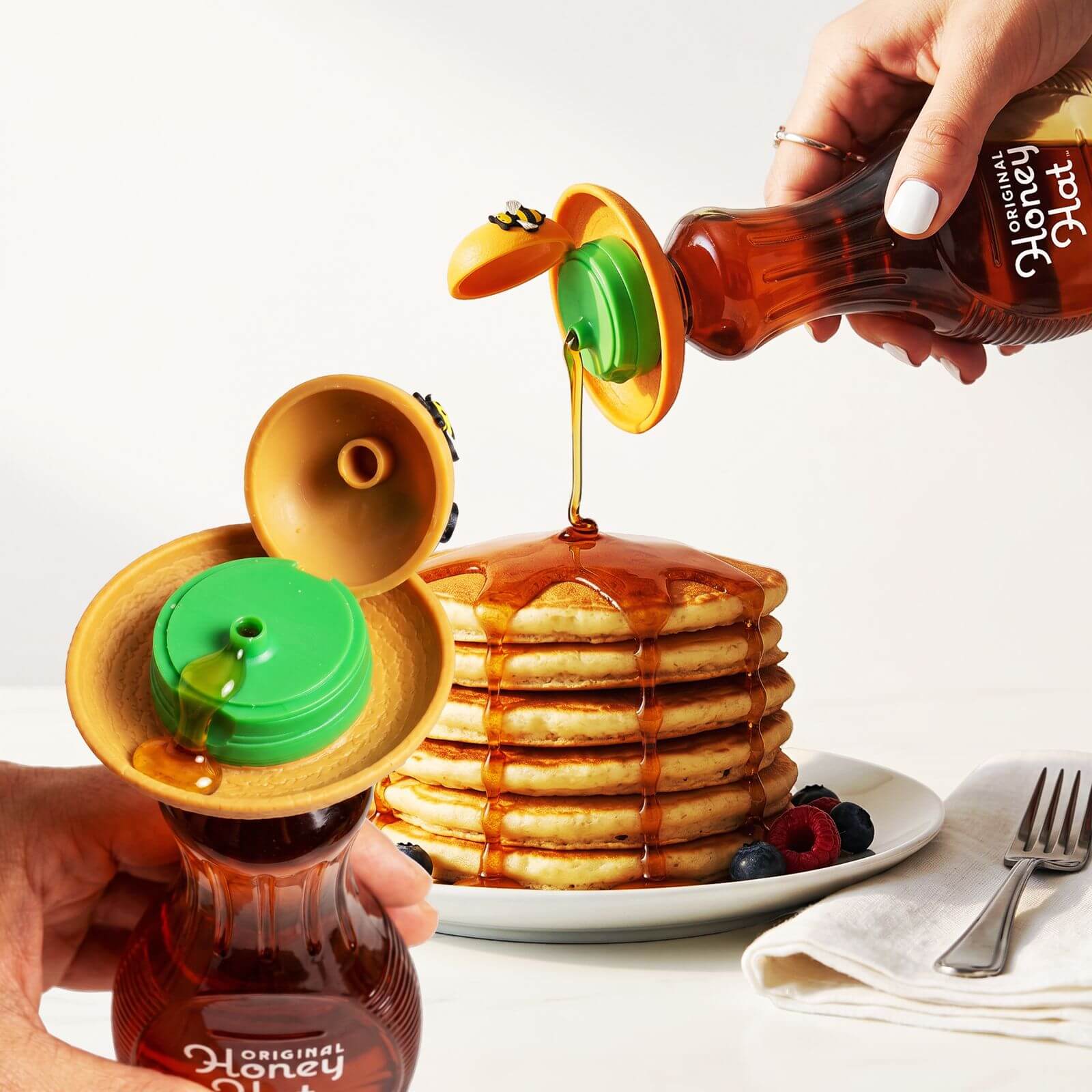 Person using the Original Honey Hat on a honey bear bottle and a syrup bottle, pouring syrup over a stack of pancakes with berries.